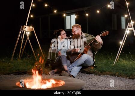 Junges Paar, kaukasische Frau und rothaarige bärtige Mann, auf einem nächtlichen Picknick mit einer Gitarre und marshmello haben eine gute Zeit Stockfoto