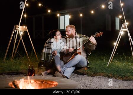 Junges Paar, kaukasische Frau und rothaarige bärtige Mann, auf einem nächtlichen Picknick mit einer Gitarre und marshmello haben eine gute Zeit Stockfoto