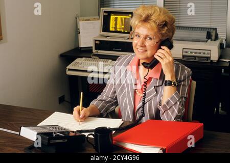 EINE GESCHÄFTSFRAU AUS DEN 1990ER JAHREN AM SCHREIBTISCH, DIE AM TELEFON SCHREIBT UND MULTITASKING ANSPRICHT BLICK AUF DIE KAMERA - KO3405 NET002 HARS JOBS KOMMUNIKATION KOPIE RAUM HALBE LÄNGE DAMEN COMPUTER PERSONEN BERUF GESCHÄFTSFRAU BLICKKONTAKT GESCHICKLICHKEIT BERUF GLÜCK FÄHIGKEITEN FRÖHLICH HIGH-WINKEL-KUNDENSERVICE KARRIERE FÜHRUNG ARBEIT BESCHÄFTIGUNG BERUFE HANDYS LÄCHELT MULTITASKING TELEFONE FREUDIGE GESCHÄFTSFRAUEN MITARBEITER PANORAMA MID-ADULT MID-ADULT FRAU KAUKASISCHEN ETHNIZITÄT COMPUTING LABORING ALTMODISCH Stockfoto