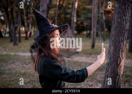 Junge nette schöne Frau in dunklem Kleid und Hexenhut macht ein Selfie für soziale Netzwerke. Halloween Party Kostüm. Wald, Park mit Herbstbäumen Stockfoto