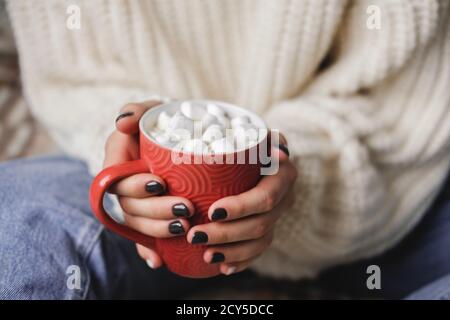 Die junge Frau sitzt auf Karos in kuschelig gestricktem weißen Wollpullover und hält eine Tasse Kakao mit Marshmallows in den Händen. Hygge Neujahr, gemütliche Weihnachten, Stockfoto
