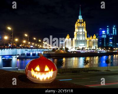 Halloween Kürbis auf dem Hintergrund der Nachtstadt. Mehrstöckige Gebäude und Wolkenkratzer. Verschwommene farbige Lichter werden im Wasser reflektiert. Nacht Stockfoto