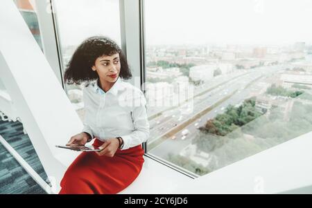 Eine schöne afroamerikanische Unternehmerin in einer weißen Bluse und Roter Rock hält einen digitalen Tablet-pc während des Sitzens Drinnen auf der Bank auf t Stockfoto
