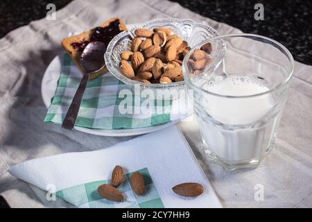 Nahaufnahme von oben auf ein Glas Milch, eine Schüssel mit Mandeln und einige verstreut auf dem Tisch und ein Toast mit Marmelade Stockfoto