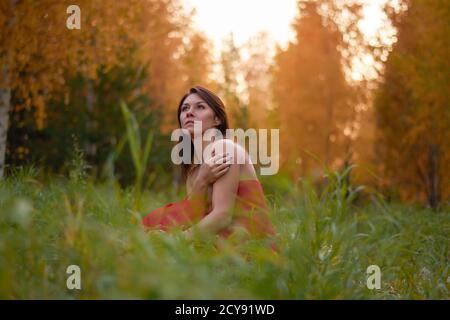 Schöne junge Frau in rotem Kleid sitzt im Herbstwald. Stockfoto