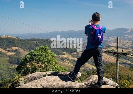 Ein Mann, der auf einem großen Felsen steht und Bilder von der bergigen Serra do Mar (Sea Ridge) Berglandschaft auf dem Gipfel des Pedra da Macela Wahrzeichen macht. Stockfoto