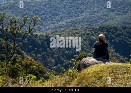 Ein junger Mann sitzt auf einem Stein in der Nähe Pedra da Macela Wahrzeichen genießen die bergige Aussicht auf Serra do Mar (Sea Ridge) Landschaft. Stockfoto