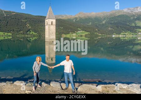Ein paar Touristen posieren vor dem alten Glockenturm des Graun Vinschgau, der sich perfekt im stillen Wasser des Reschsees, Südtirol, widerspiegelt. Stockfoto