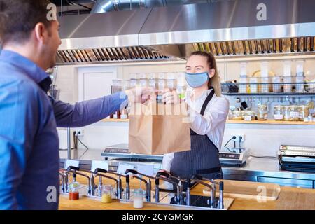 Glückliche Kellnerin trägt eine Gesichtsmaske und serviert dem Gast am Tresen im kleinen Familienrestaurant Essen zum Mitnehmen. Stockfoto