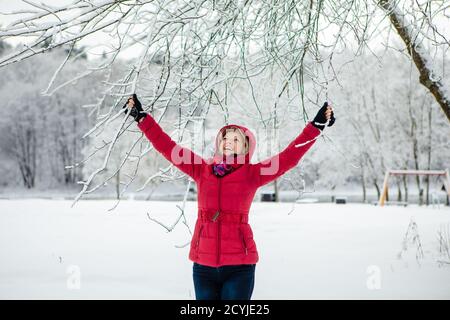 Kaukasische weiße Frau mit roter Jacke genießen verspielt am verschneiten Tag mit verschwommenem weißen Wald Hintergrund.Weihnachtszeit. Glück. Schneefälle. Stockfoto
