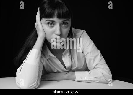 Studioportrait einer hübschen Brünette in einem weißen Hemd, auf einem weißen Tisch gelehnt, vor einem schlichten schwarzen Hintergrund, auf die Kamera schauend Stockfoto