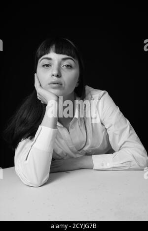 Studioportrait einer hübschen Brünette in einem weißen Hemd, auf einem weißen Tisch gelehnt, vor einem schlichten schwarzen Hintergrund, auf die Kamera schauend Stockfoto