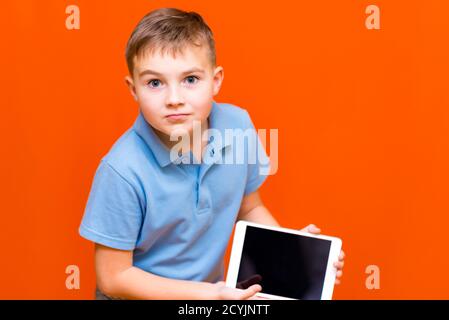 Porträt des europäischen Schuljungen zeigt Tablet und blaues T-Shirt auf Orange Studio background.cheerfully denkt etwas nach und zeigt etwas auf leer ta Stockfoto