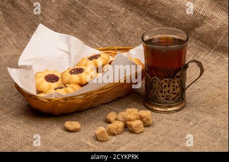 Hausgemachte Gebäck-Kekse mit Marmelade und einem facettierten Glas Tee in einem Vintage-Cup-Halter auf einem Hintergrund von homespun Stoff mit einer rauen Textur, close-up, Stockfoto