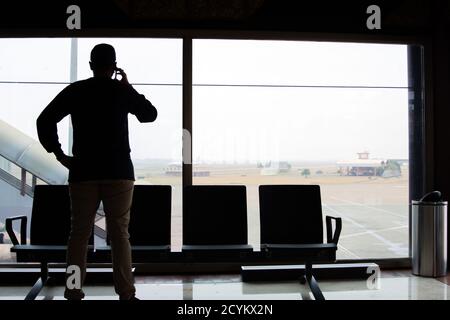 Die Silhouette der Rückansicht eines Mannes macht Ein Telefonanruf, während Sie vor einem Glas stehen In einem Wartezimmer am Flughafen Stockfoto