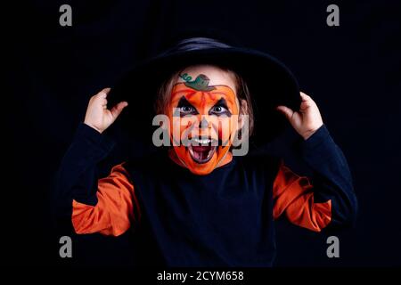 Ein Kind mit Kürbis Make-up in einem schwarzen Hut hält seine Hände an den Hut und lacht laut auf schwarzem Hintergrund, isoliert, Stockfoto