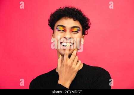 Glücklicher junger Mann trägt regenbogenfarbenen Lidschatten und Lächeln Gesicht Nagelfarbe. Homosexuell männlich lächelnd mit Hand auf Gesicht vor rotem Hintergrund. Stockfoto