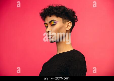 Hübscher Mann in schwarzem T-Shirt starrt in die Kamera. Androgynen Mann in schwarzem Outfit und regenbogenfarbenen Lidschatten vor rotem Hintergrund. Stockfoto