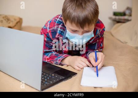 Teenager liegt auf dem Bett mit Laptop und macht Hausaufgaben. Kind Schulkind lernt während Quarantäne von Coronavirus. Student in schützende medizinische Maske engag Stockfoto