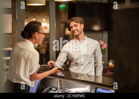 Eine Dame an der Rezeption, die die kontaktlose Bezahlung eines Kunden übernimmt, der im Hotel eincheckt, verwendet sein Smartphone, um zu bezahlen. Stockfoto