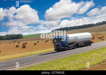 White Tanker fahren entlang der Asphaltstraße um das gelb geerntete Weizenfeld in der Landschaft. Blauer Himmel mit weißen Wolken. Stockfoto