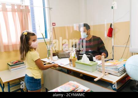 Schüler mit einer Maske, die Hausaufgaben an den Lehrer übergibt Durch einen Bildschirm Stockfoto