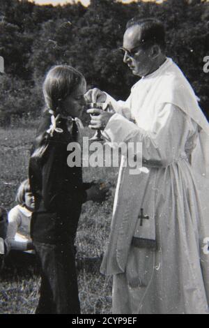 Schöne Vintage-Schwarz-Weiß-Fotografie aus den 1970er Jahren, bei der ein junges Mädchen während der Messe die heilige Kommunion von einem Priester empfängt. Stockfoto