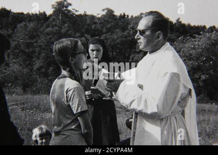 Schöne Vintage-Schwarz-Weiß-Fotografie aus den 1970er Jahren, bei der ein junges Mädchen während der Messe die heilige Kommunion von einem Priester empfängt. Stockfoto