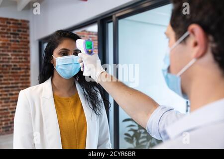 Mischrasse Frau und kaukasischen Mann arbeiten in einem lässigen Büro, tragen Gesichtsmasken, ein Mann nimmt ihre Temperatur. Soziale Distanzierung am Arbeitsplatz du Stockfoto