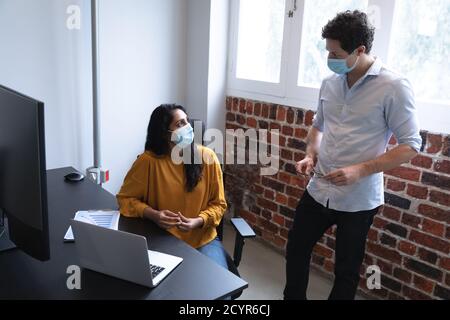 Gemischte Rasse Frau und kaukasischen Mann arbeiten in einem lässigen Büro, tragen Gesichtsmasken und sprechen. Soziale Distanzierung am Arbeitsplatz während des Coronavirus C Stockfoto