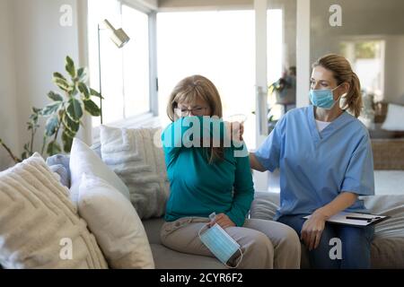 Kaukasische Frau, die zu Hause von einer kaukasischen Krankenschwester besucht wird, bedeckt den Mund beim Husten, die Krankenschwester trägt Gesichtsmaske. Medizinische Versorgung zu Hause du Stockfoto