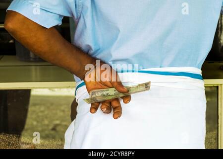 Mann mit Lungi an einem Ladenschalter der Main Road mit 100 Rupien-Note in dieser geschäftigen Stadt im Wayanad-Distrikt; Kalpetta, Wayanad, Kerala, Indien Stockfoto