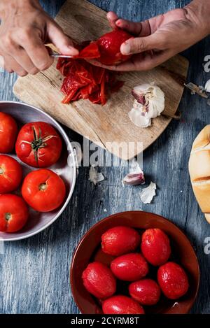 Hochwinkelansicht eines jungen kaukasischen Mannes, der einige gebrühte Tomaten auf einem grauen rustikalen Küchentisch schält, neben einem Metallteller mit einigen rohen Tomaten, ein Stockfoto