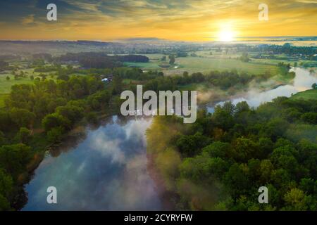 Ein gewundener Fluss im Nebel zwischen den Bäumen dahinter Das Dorf in der Morgendämmerung in den Strahlen der warmen So Stockfoto