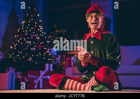 In voller Länge Foto von verrückt elf sitzen Boden halten smart Telefon im Haus drinnen mit weihnachten Weihnachten noel Abendbeleuchtung Stockfoto