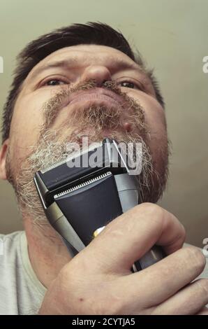 Ein erwachsener Mann rasiert sich mit einem elektrischen Rasiermesser. Ungewöhnliches Porträt eines bärtigen Mannes mit grauem Haar. Grimassen vor der Kamera Stockfoto