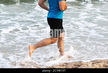 Seitenansicht eines Mannes, der barfuß im Wasser am Rande des Strandes läuft und planscht. Stockfoto