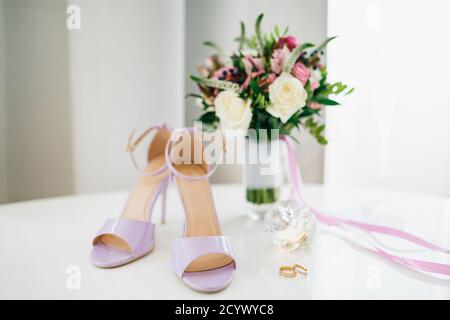 Lila Brautsandalen auf dem Tisch mit den Eheringen der Braut und des Bräutigams und einem Blumenstrauß in einer Vase. Stockfoto