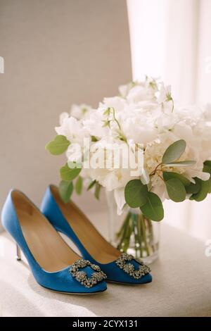 Blaue Damenschuhe mit einer Schnallenspitze auf einer weißen Oberfläche mit einem Bouquet von Pfingstrosen in einer Vase. Stockfoto