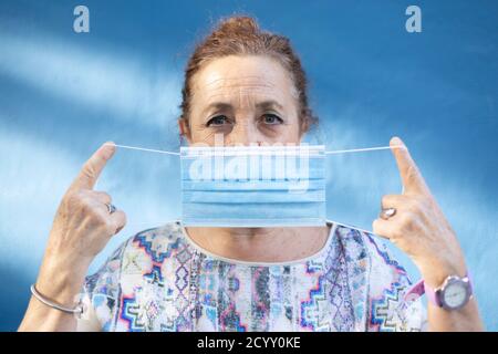 Ältere Frau, die eine medizinische Maske auf ihr Gesicht legt. Sie steht neben einer blauen Wand. Stockfoto