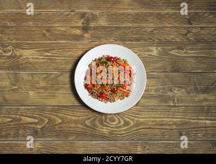 Gemüsesalat mit warmem Buchweizen, Tomaten, Paprika, Zwiebeln, Olivenöl und verschiedenen Kräutern auf einem weißen Teller auf Holzgrund, Draufsicht w Stockfoto