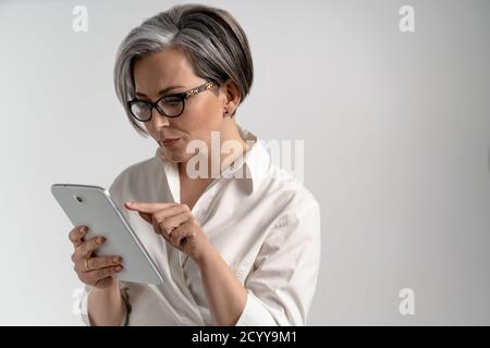 Elegante Frau mit Brille hält Tablet und drückt ihren Finger. Reife Frau mit grauen Haaren in weißem Hemd auf einem isolierten Hintergrund. Hohe Qualität Stockfoto