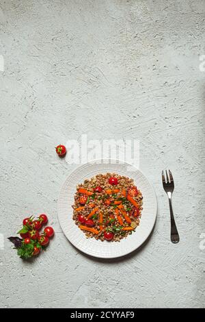 Gemüsesalat mit warmem Buchweizen, Tomaten, Paprika, Zwiebeln, Olivenöl und verschiedenen Kräutern auf einem weißen Teller auf grauem Hintergrund, Draufsicht mit Stockfoto