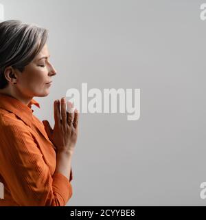 Schöne Erwachsene Frau steht im Profil mit gefalteten Handflächen und geschlossenen Augen. Frau mit grauen Haaren auf einem isolierten Hintergrund im Studio. Gebetskonzept Stockfoto