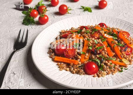 Gesunder vegetarischer Salat mit warmem Buchweizen, Tomaten, Paprika, Zwiebeln, Olivenöl und verschiedenen Kräutern auf einem weißen Teller auf grauem Grund Stockfoto