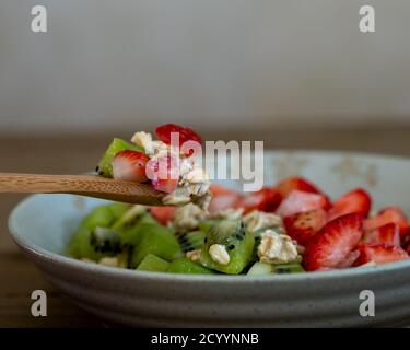 Holzlöffel, der eine Portion Haferflocken von einem Bräunungsteller aufsammelt, garniert mit gewürfelten Erdbeeren und Kiwi auf einem Holztisch, ganzer Teller, zentriert, Seite Stockfoto