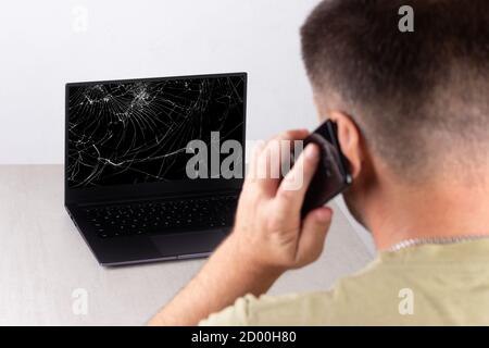 Ein junger Mann, der auf einem Mobiltelefon vor einem Laptop mit einem defekten, geknackten Bildschirm spricht, das Konzept der Kontaktaufnahme mit dem technischen Support oder Computer Repa Stockfoto