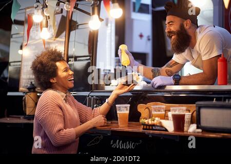 Der Mitarbeiter von Food Truck gibt ein frisch zubereitetes Sanwich Eine glückliche junge afro amerikanische Frau Stockfoto