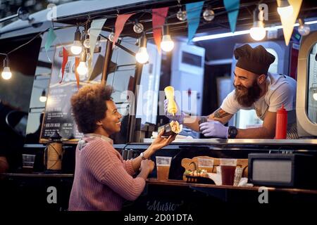 Der Mitarbeiter von Food Truck gibt ein frisch zubereitetes Sanwich Eine glückliche junge afro amerikanische Frau Stockfoto