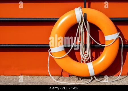An orange life ring hangs on a hook on a black fence in front of an orange background surface. The life preserver has white ropes attached. Stockfoto